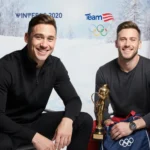 Adam Rippon smiles while resting arm on microphone stand with Chase Cain nearby holding trophy against snow backdrop Team USA