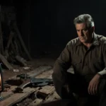 Alec Baldwin sitting on dusty Rust set with flickering lantern and broken gun nearby melancholy mood