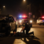Trooper kneeling beside lifeless driver with mangled car in front and police lights overhead