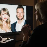Andrea Swift sits with warm glow from screen showing Taylor Swift and Travis Kelce while a football key rests nearby
