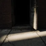 Apartment doorway ajar revealing sliver of decay with light spilling onto cracked sidewalk and dim interior