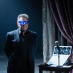 Barry Manilow standing before dim stage with blue glow on his face and a medical chart and MRI scan near the mic