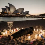 Lone figure lying on Bondi Beach sand with candles and menorahs under dawn light near Opera House