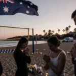 Group of mournful faces standing with tears and clasped hands under an Australian flag at dusk on Bondi Beach