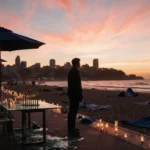 Lone figure standing near shattered table gazing at Bondi Beach sunset with candles and abandoned umbrellas