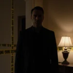 Nick Reiner standing alone in foyer with faint lamp glow and police tape on walls