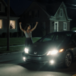 Young woman threatens with arms raised beside Honda Civic with headlights blazing toward camera and suburban house looming
