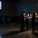 Students gathering and looking shocked with glowing phones in a dim gym while a screen shows a live stream.