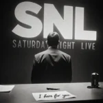 Chevy Chase standing alone in front of a dimly lit desk with a broken pen and faded SNL logo behind him