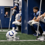 Quarterback sits on NFL field sidelines with abandoned helmet and teammates using ice packs and crutches
