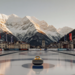 Gold curling stone resting on pedestal with sunset-lit Dolomite mountainside and town buildings