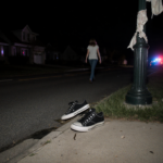 Shoes lie abandoned on dimly lit street with torn fabric hanging and distant police lights flicker
