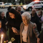 Family members holding hands around a makeshift memorial with warm lighting and police tape in background