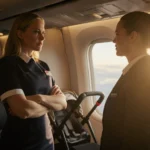 Amy Poehler defies airline crew member with arms while a baby stroller sits behind her in first‑class sky hinting turbulence.