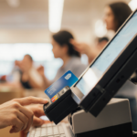 Clerk reaching to remove a card skimmer with a retail terminal screen and blurred cashier and customer hands in background