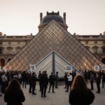 Museum staff protesting with signs in front of the Louvre glass pyramid at dusk with blurred tourists and guards.