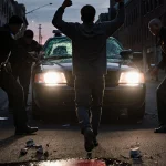 Teenager raising arms with police officers and flashing police car headlights in a dark Philadelphia street