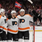 Carter Hart beams while holding his stick and being high‑fived by teammates with a cheering crowd of Flyers fans behind him