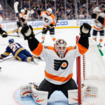 Flyers goalie celebrates win with arms raised and teammates high‑fiving around him after 5-2 victory while Sabres slump on ic