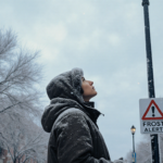 Bundled‑up person gazing up at the winter cold front with frost‑covered street and faint Frost Alert sign in Philadelphia.