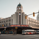 Historic Greyhound Depot entrance with restored sign and buses parked in front near bustling city street and skyline.