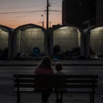 Mother and child sitting on bench with heads bowed surrounded by rows of glowing shelters at dusk in Philadelphia