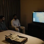 Father and son sit in room with newspaper and open suitcase on bed showing travel itinerary near TV.