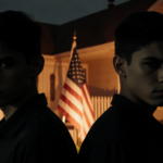 Twins standing back-to-back with stern faces in front of a dimly lit suburban home and a faint American flag silhouette.