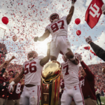 Hoosier football players lifting teammates onto shoulders with confetti and a giant trophy near fans holding Indiana flags