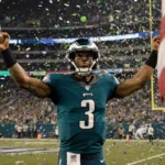 Jalen Hurts raising arms with confetti and a giant American flag in a packed Philadelphia Eagles stadium.