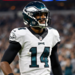 Jalen Hurts stands on sideline with Philadelphia Eagles helmet and pads while fans blur in background.