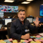 Jimmy Kimmel sits at his desk holding a microphone with colorful notes and a TV screen showing the ABC logo in a bustling new