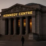 Gold lettering on Kennedy Center façade glows with dusk light near Washington DC skyline and a lone passerby walks away.