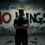 Kevin Krebs stands with arms crossed against graffiti wall with No Kings banner and discarded guns nearby and guilty