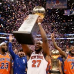 Knicks player holding NBA Cup trophy with confetti exploding and fans in orange and blue jerseys at Madison Square Garden.