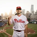 Kyle Backhus stands triumphantly baseball stadium with Phillies jersey and bat over shoulder near Philadelphia skyline sunlit