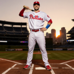 Kyle Schwarber stands at home plate holding a $150M contract with a bat and Phillies jersey shining under sunset lights