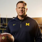 Kyle Whittingham standing with a football at his feet and sunset glow over the Maize and Blue Michigan field
