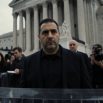 Luigi Mangione walking out of courthouse with officers and reporters behind plastic barrier.