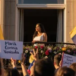 María Corina Machado standing on balcony overlooking supporters with Venezuelan flags and flowers.