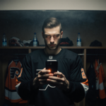 Matvei Michkov reviewing game footage with phone glow and foggy mirror in dim Flyers locker room