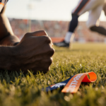 Micah Parsons rests a clenched fist on field grass with a torn ACL ribbon nearby and a blurred Denver player in motion
