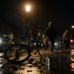 Shattered bike lies abandoned on wet pavement with streetlight glow and faint police tape in background
