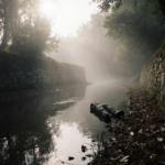 Body lies beside weathered stone wall in misty creek with sunlight filtering through trees.