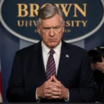 Male official speaking at podium with navy American flag and Brown University logo beside him under harsh spotlight