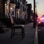 Police cruiser stands with flashing lights and abandoned chair on stoop amid dim dusk street.