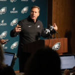 Eagles coordinator gesticulating passionately at podium with reporters and microphones around