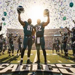 Phillips and Carter raise Eagles helmets in victory with teammates cheering under golden light and confetti.
