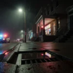 Streetlight casts long shadows on wet pavement in Philadelphia with bloodstain and flashing police cruiser nearby