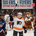Hockey player smiles holding new stick with Flyers logo and surrounded by Sixers Joel Embiid Tobias Harris mascots.